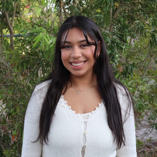 Smiling young woman with long dark hair wearing a white cardigan, standing in front of leafy bushes.