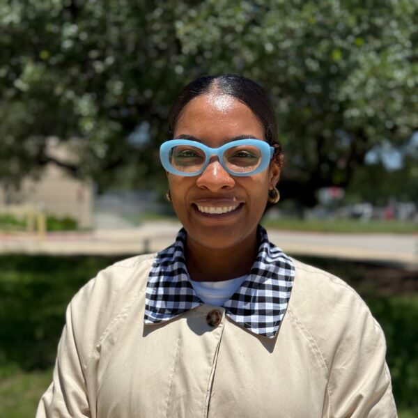 Close-up selfie of a smiling person outdoors, wearing light beige coat, blue sunglasses, and a checkered shirt in sunny daylight.