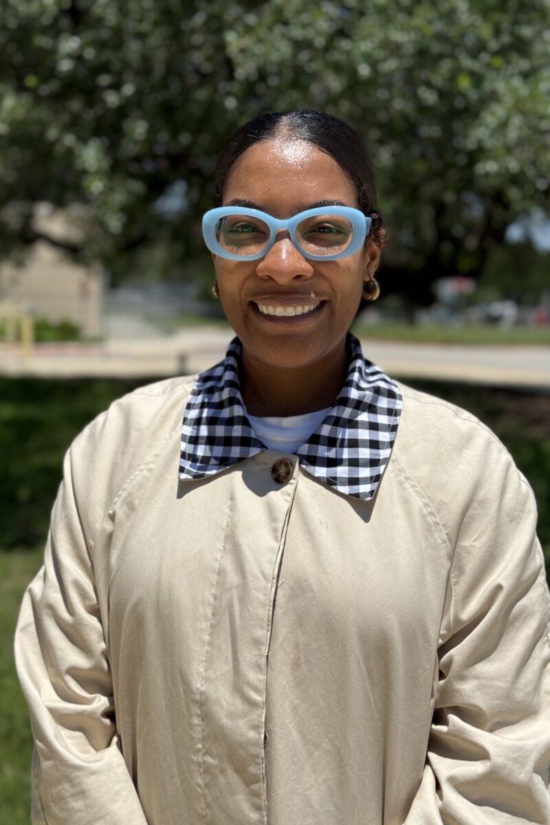 Portrait of a smiling woman wearing a light beige trench coat and blue oversized glasses outdoors on a sunny day with trees behind her.